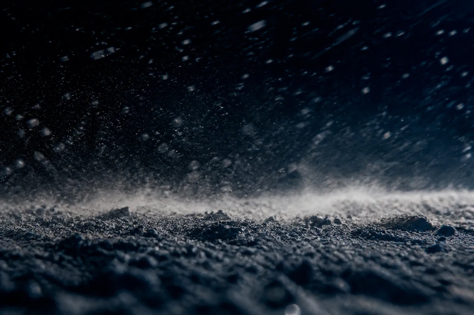 A close, almost abstract view of pale alkali dust and grit driven by wind across dark ground at night.