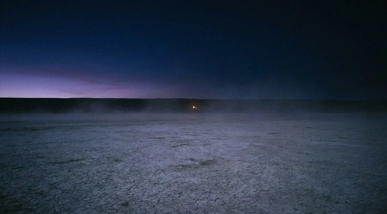 A vast cracked alkali flat stretches to a distant horizon under a deep indigo sky at late dusk. A single point of amber light shines in the extreme distance.
