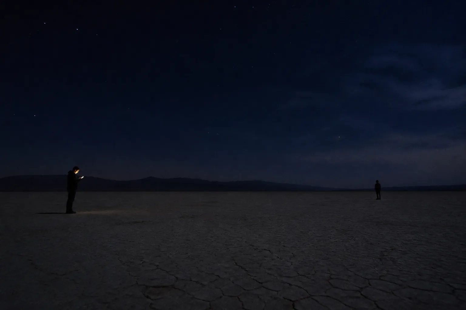 Two small figures stand far apart on a flat desert playa at night, under stars. One is lit by the warm amber glow of the phone screen they are holding.